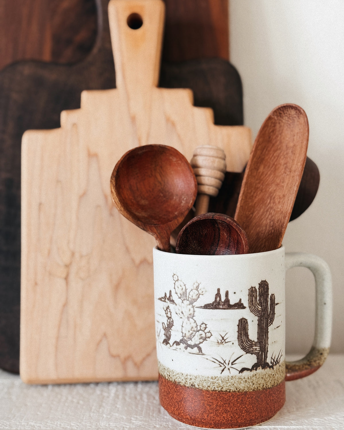 Ceramic mug with desert design containing wooden spoons, against a neutral background
