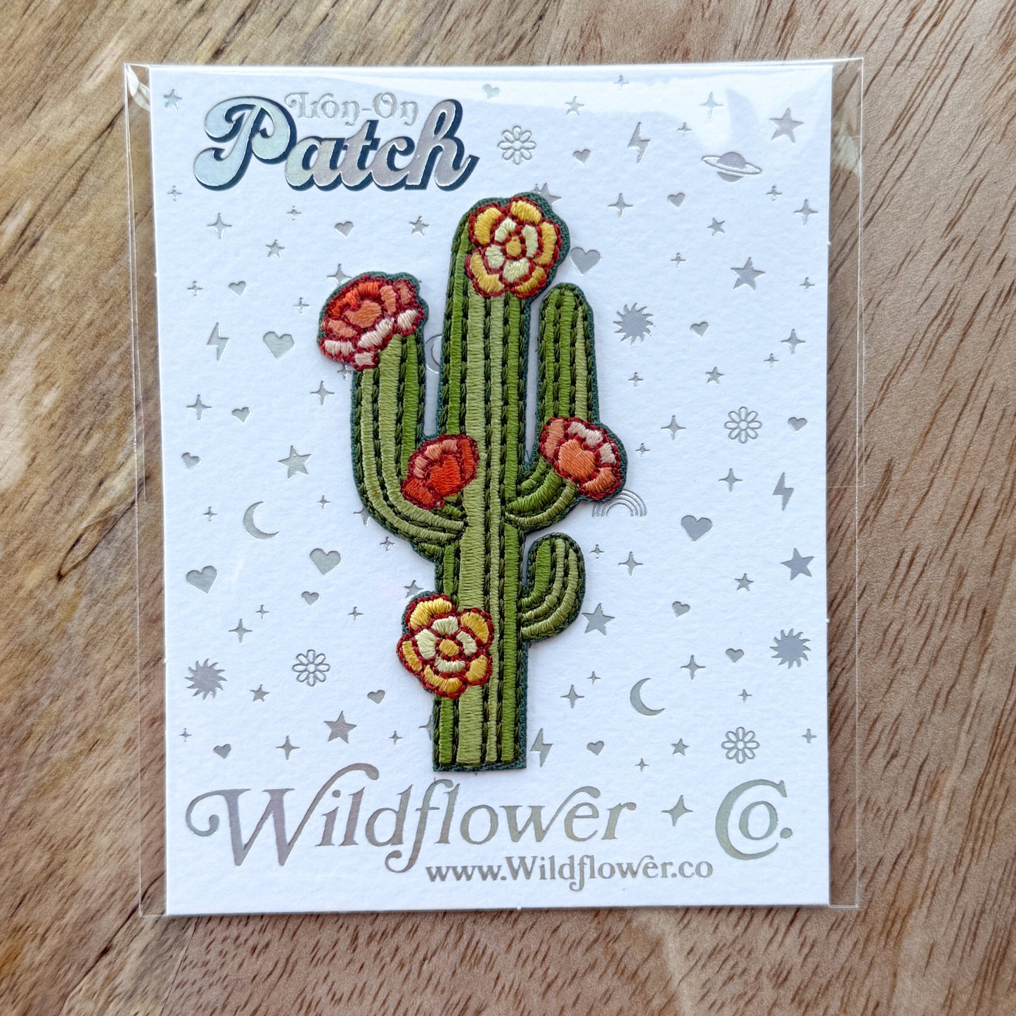 Embroidered patch of a cactus with flowers on a Wildflower Co. card, placed on a wooden surface.