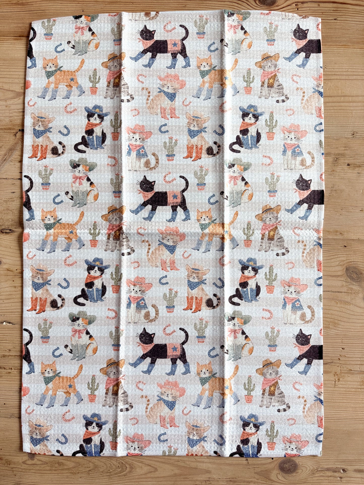 A rectangular kitchen tea towel with a waffle-weave texture, featuring a colorful print of cartoon cats in cowboy hats and cacti, laid flat on a light brown wooden surface.
