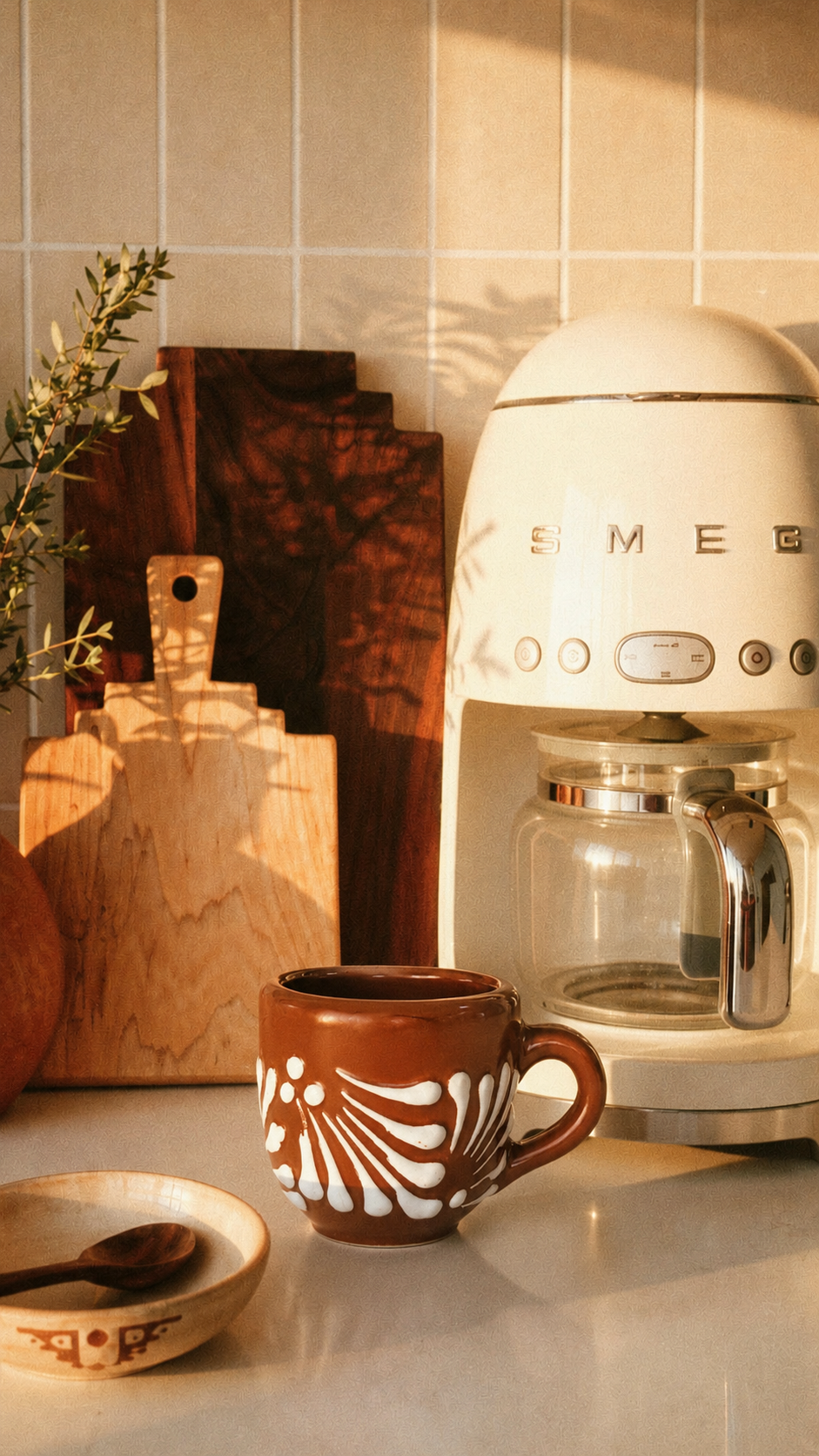 A retro-style cream-colored SMEG coffee maker sits on a kitchen counter next to a brown ceramic mug with white leaf patterns, with warm sunlight casting shadows on a tiled wall.