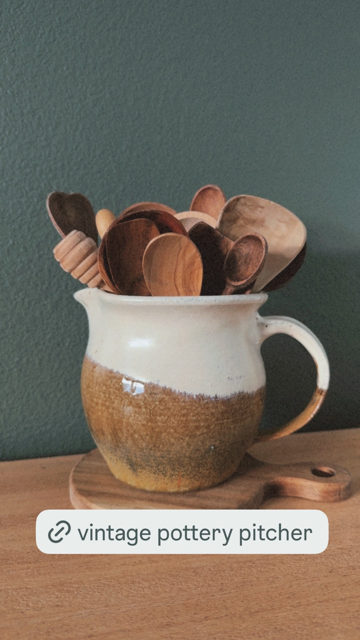 A vintage-style ceramic pitcher with a white upper body and a brown glazed lower body, filled with wooden spoons, sitting on a wooden surface against a dark green wall.