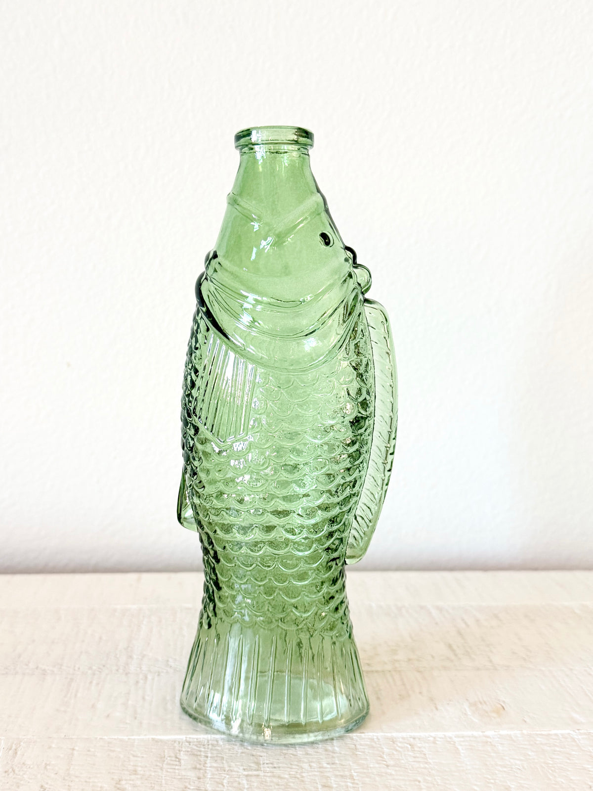 A light green glass bottle shaped like a fish, featuring a textured scale pattern and a ribbed base, standing on a light wooden surface against a white wall.