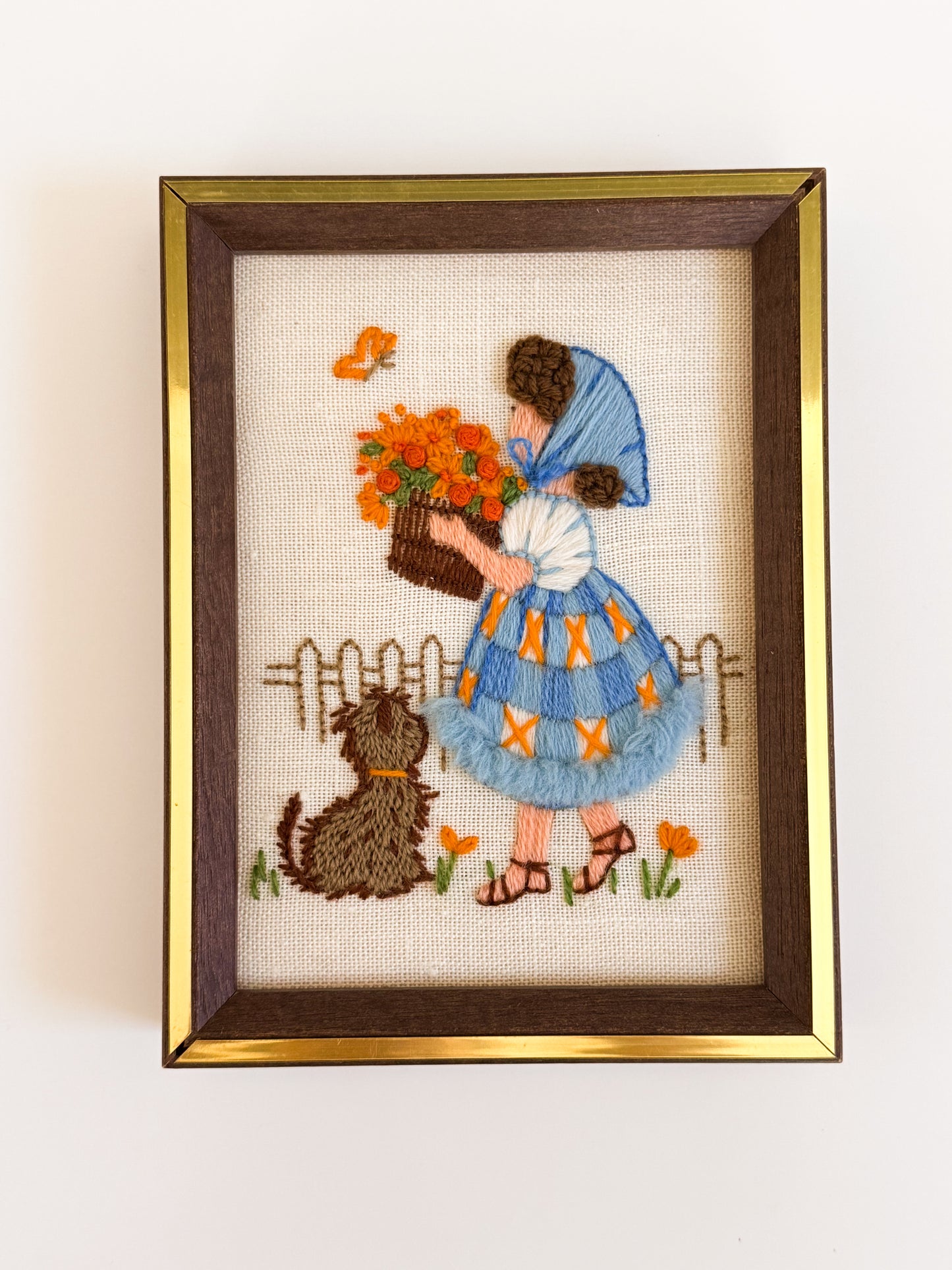 Embroidery of a girl with flowers and a dog in a gold frame on a white wall.