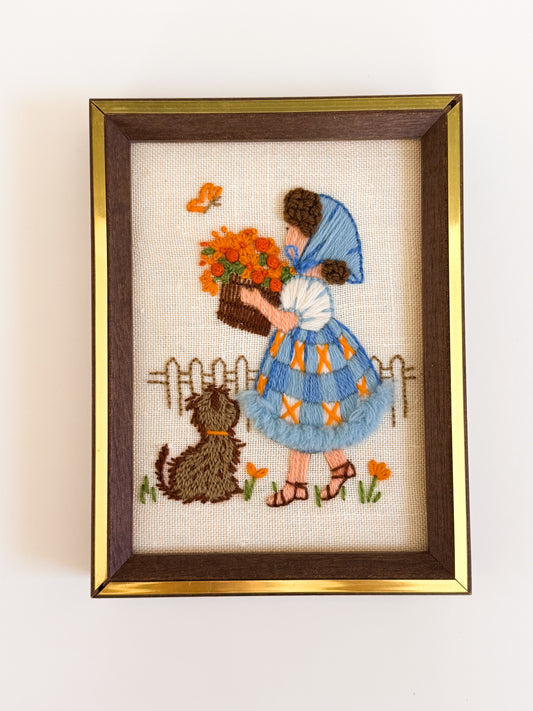 Embroidery of a girl with flowers and a dog in a gold frame on a white wall.