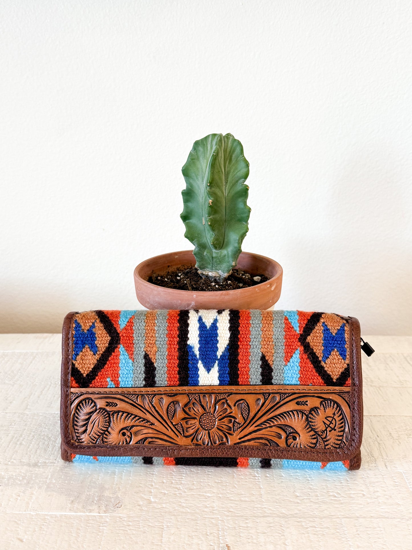 Colorful Southwestern patterned clutch with leather floral design on a white surface next to a potted plant.