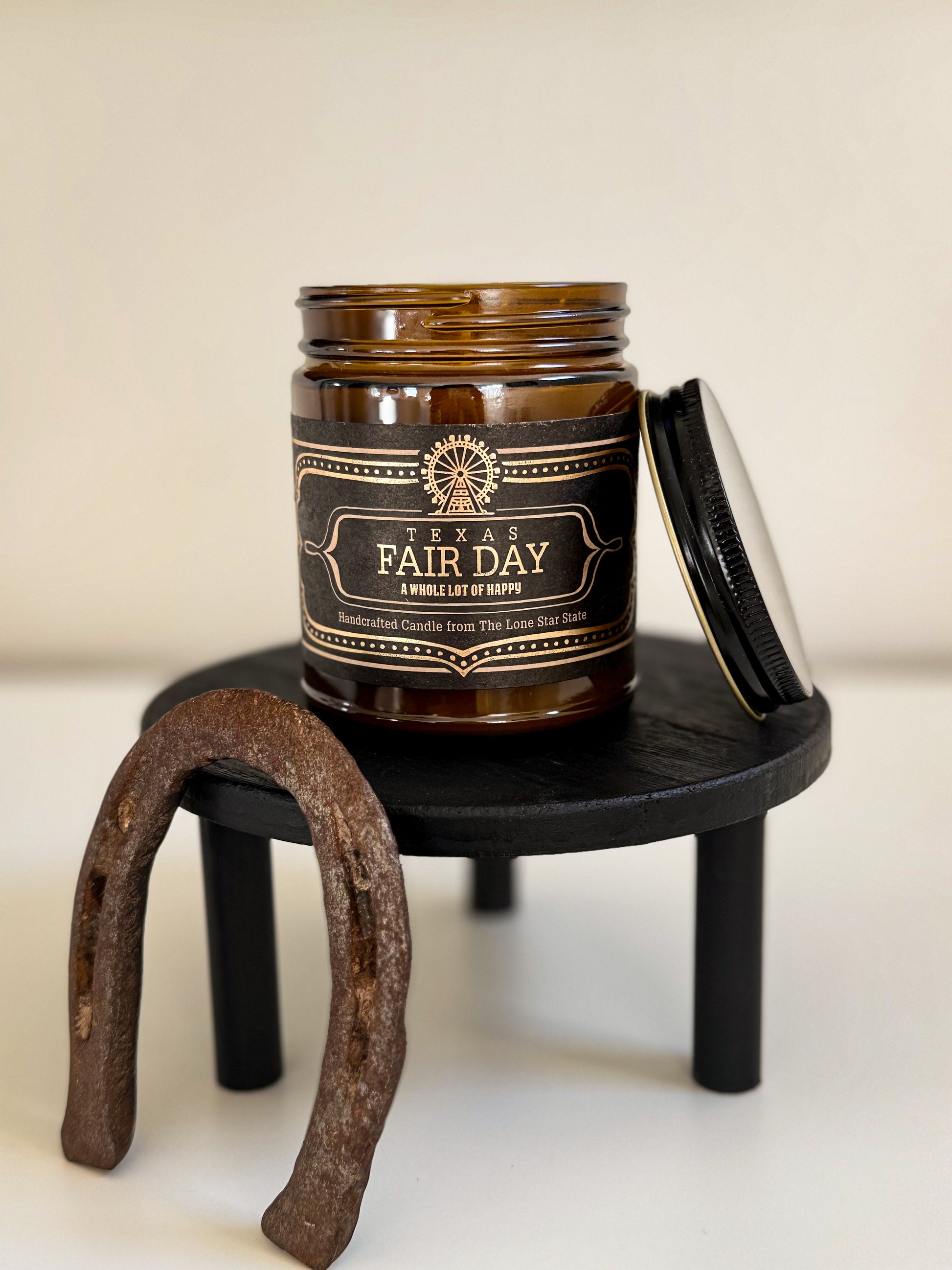 Brown candle jar labeled 'A Fair Day' on a small black stand with a horseshoe beside it.