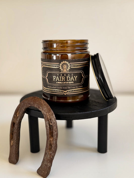 Brown candle jar labeled 'A Fair Day' on a small black stand with a horseshoe beside it.