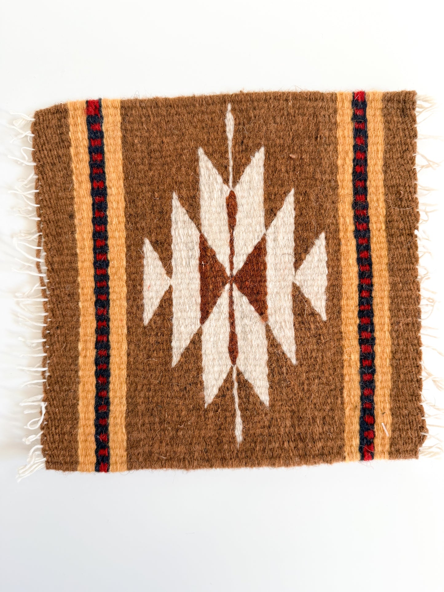 A square woven textile with a Southwestern-style geometric pattern in brown, cream, and red, featuring white fringe on the left and right edges, photographed flat against a white background.