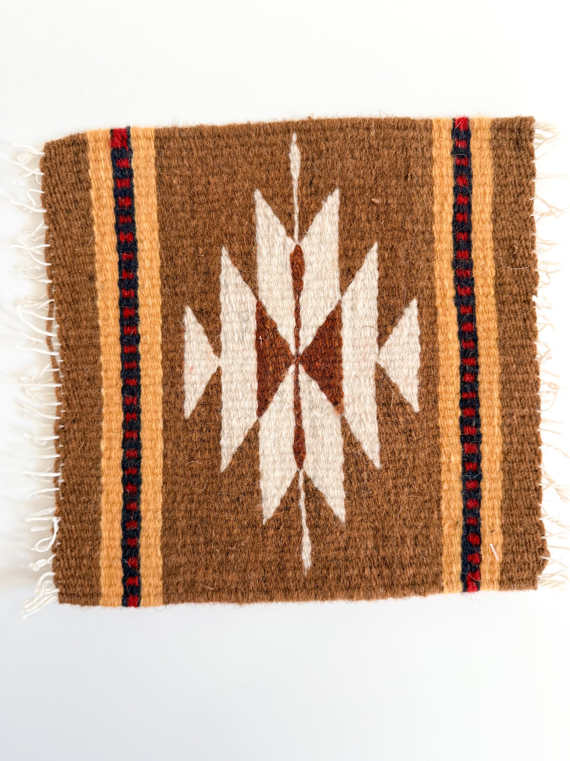 A square woven textile with a Southwestern-style geometric pattern in brown, cream, and red, featuring white fringe on the left and right edges, photographed flat against a white background.
