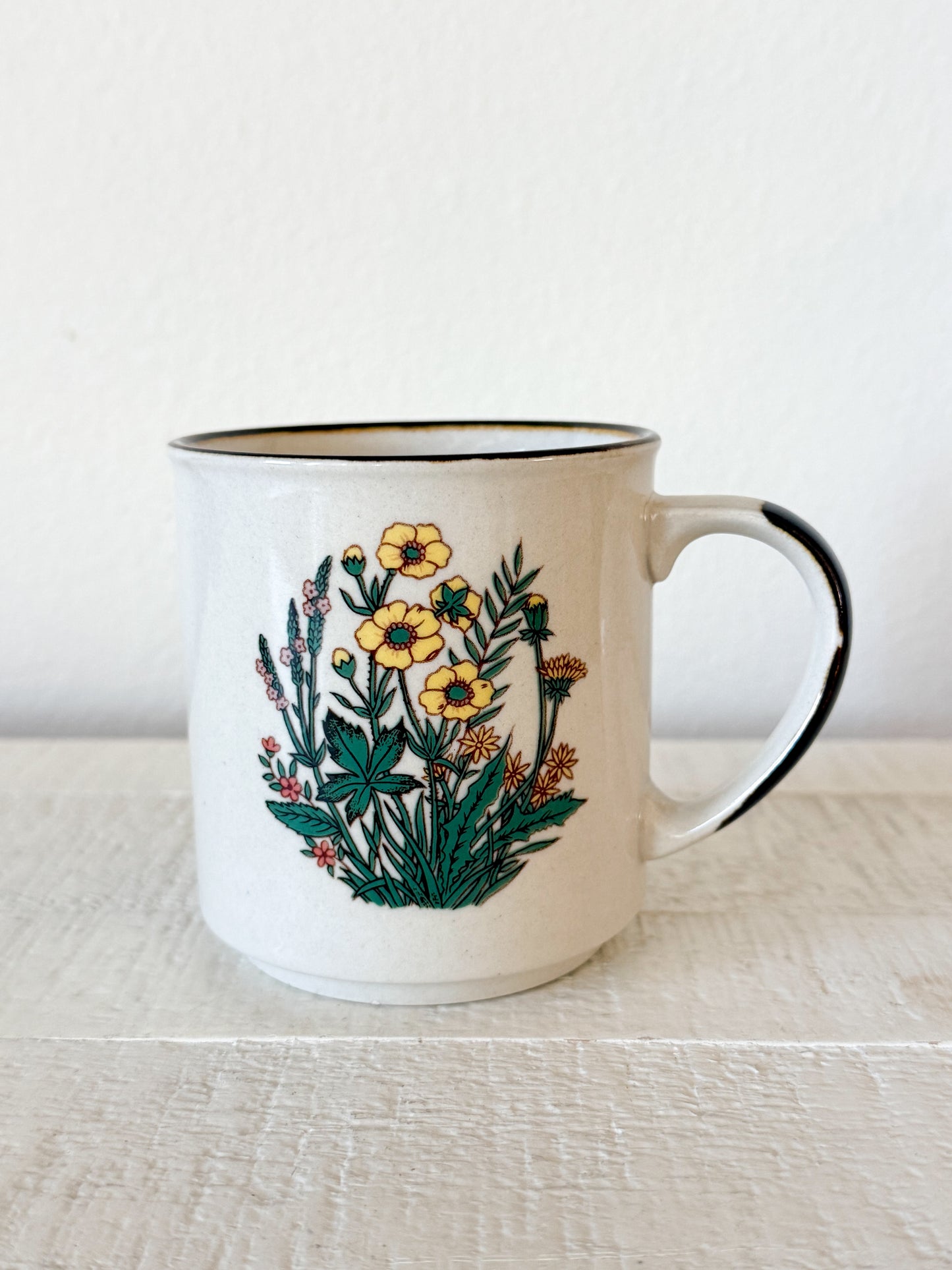 A white ceramic mug with a brown rim and handle edge, decorated with a colorful illustration of yellow wildflowers and green foliage, set on a light wooden surface against a white wall.