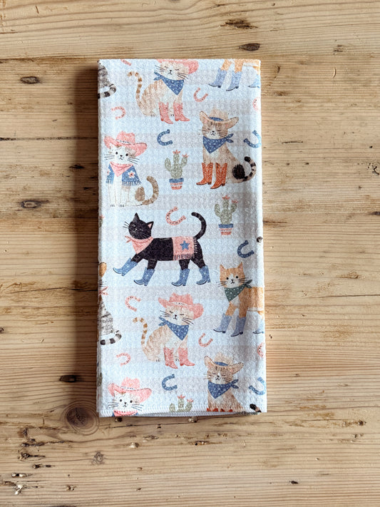 A rectangular kitchen towel with a western cat print, laid flat on a light wood surface. The towel has a white background with illustrations of various cats in cowboy hats and bandanas, along with cacti and horseshoes.