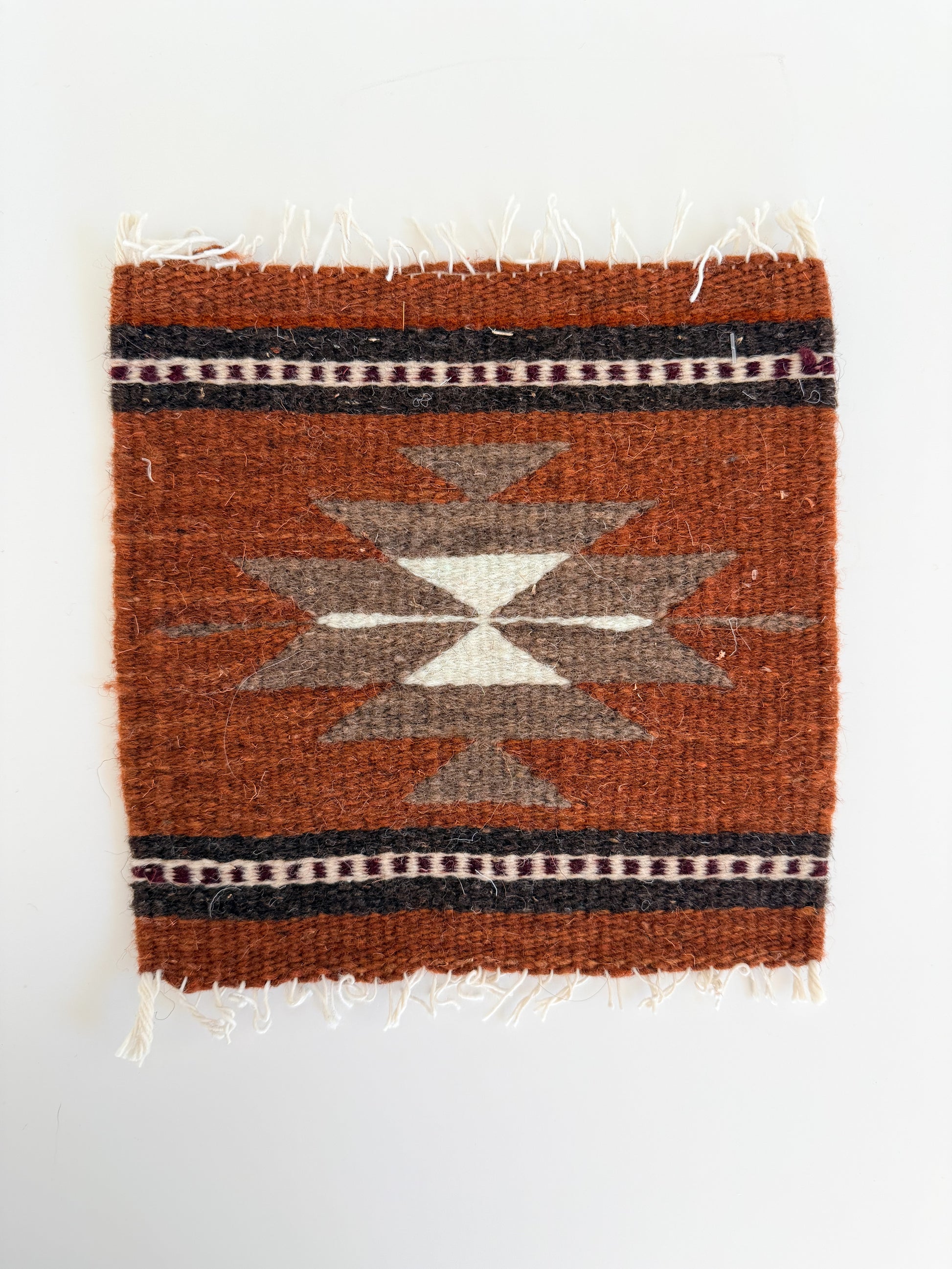 A square woven textile rug or mat with a Southwestern geometric pattern in rust orange, brown, beige, and black, shown flat against a plain white background.