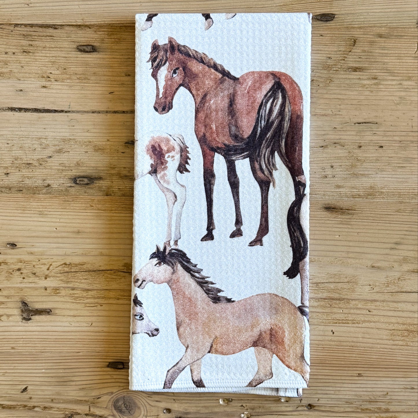 A white kitchen towel featuring a watercolor-style print of several horses, laid flat on a rustic wooden surface.