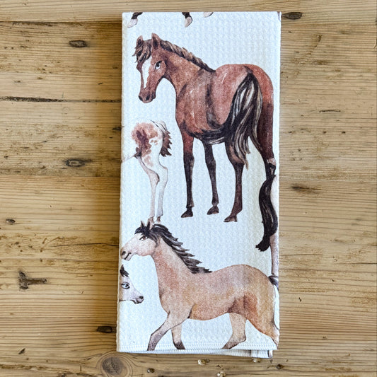 A white kitchen towel featuring a watercolor-style print of several horses, laid flat on a rustic wooden surface.