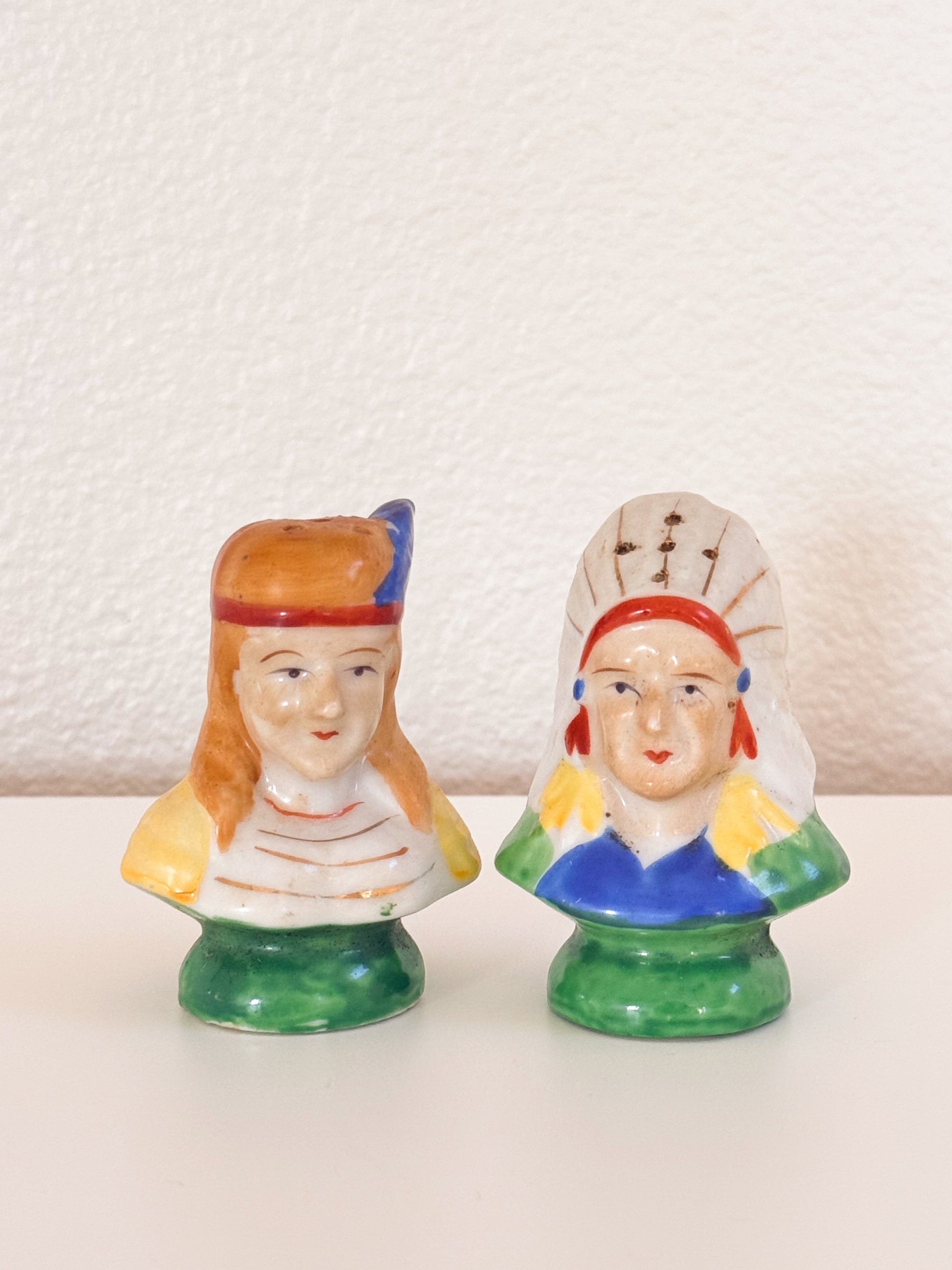 Two small ceramic figurines of Native American-style busts on a white surface against a textured off-white wall. One has orange hair and a red/blue headband with yellow and white clothing and a green base.