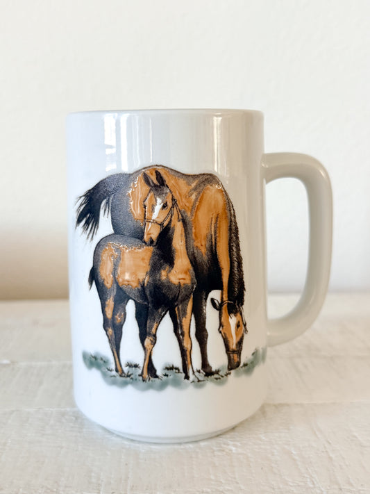 A white ceramic mug featuring a printed illustration of a brown horse and foal standing together, set against a plain light background.