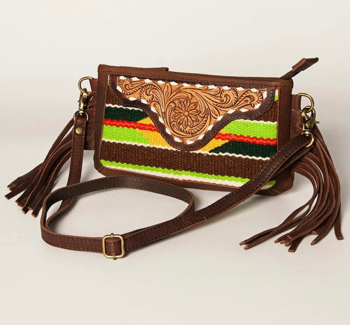 Western crossbody purse with colorful serape fabric, brown tooled leather trim, side fringe, adjustable strap, and zippered closure.